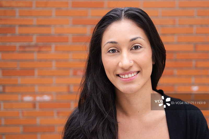 在城市环境中，站在砖墙前，笑容满面的快乐年轻拉丁裔女性图片素材