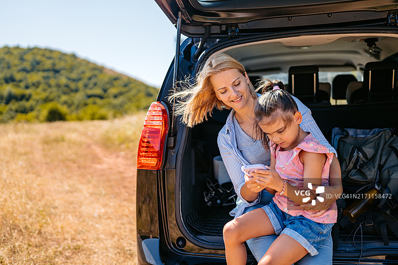 中年妇女和她的小学女儿在山上的汽车后备箱里休息并使用手机图片素材