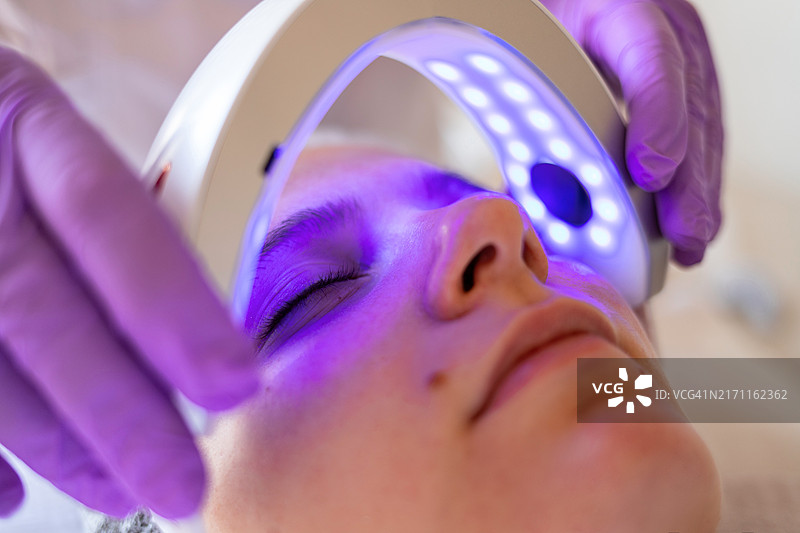 美容师手持LED蓝光灯，为年轻女性进行深层清洁和痤疮治疗图片素材