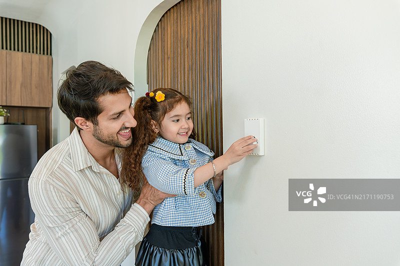 父亲和他的小女儿在家中享受简单而快乐的时刻。父亲抱着女儿，女儿伸手去调整或打开空调的开关。图片素材