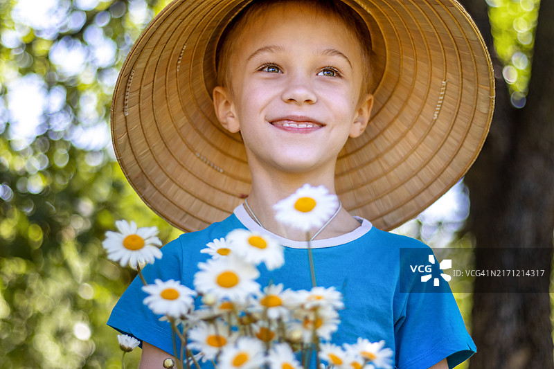 戴着越南草帽和青色衬衫，手持雏菊花束的男孩，微笑而高兴图片素材
