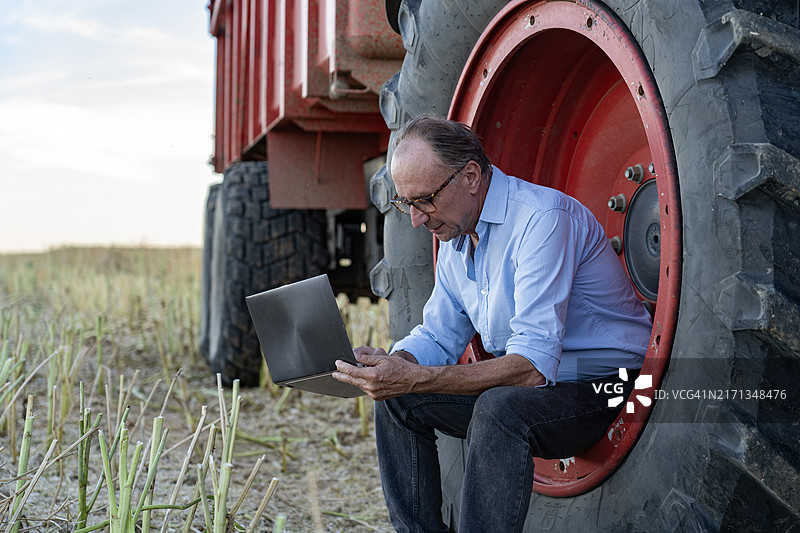 精密农业专家在耕地上使用笔记本电脑，坐在收割机的轮子上图片素材