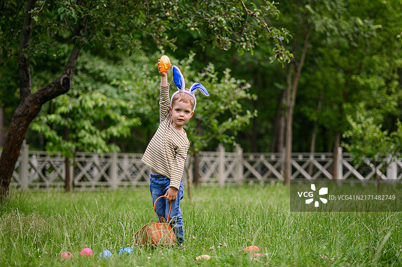 参加复活节彩蛋狩猎的儿童，戴着兔子耳朵和条纹衬衫，快乐地从草地上捡起复活节彩蛋并放入篮子图片素材