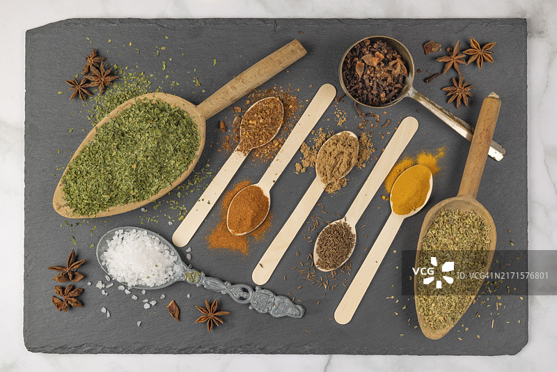 花岗岩板上不同勺子上的各种香料：牛至、海盐、辣椒粉、丁香、香菜、小茴香、姜黄，美食摄影图片素材