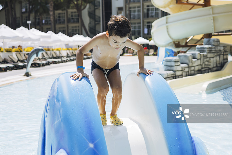 一位年轻男孩在阳光明媚的度假泳池中，沿着一个色彩鲜艳的滑水道滑下，享受暑假，附近有棕榈树图片素材