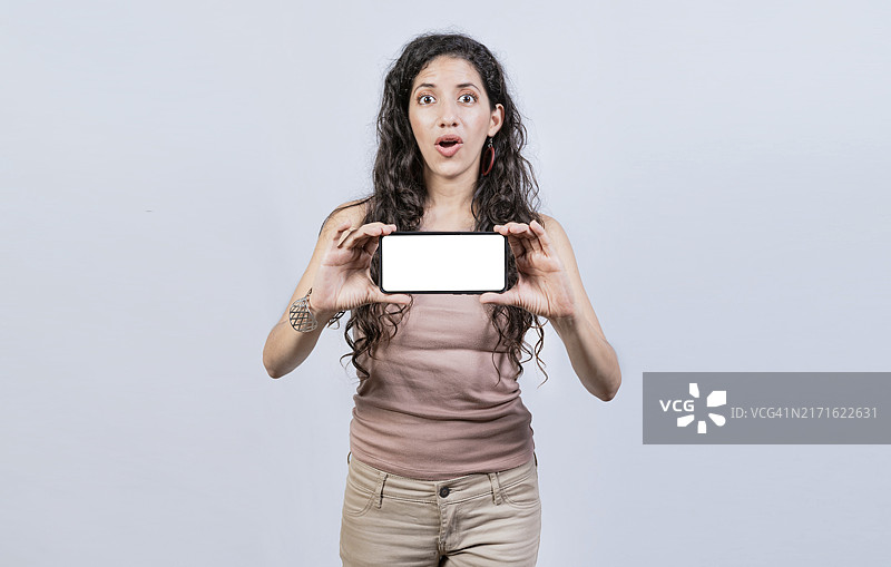 惊讶的女孩展示智能手机的空白屏幕，展示手机横屏的拉丁女孩，惊讶的年轻女子在手机上展示广告图片素材