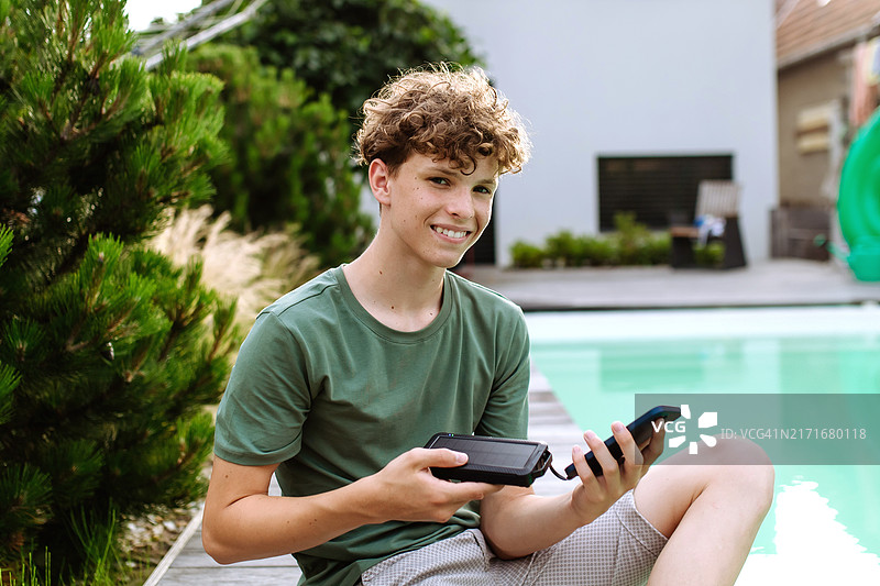 坐在游泳池边的户外，使用智能手机太阳能充电器的青少年男孩图片素材