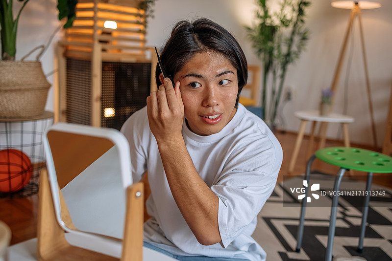 年轻的亚洲男子在家中镜子前修剪眉毛，进行自我美容护理。图片素材