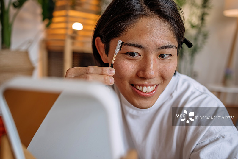 年轻的亚洲男子在家中镜子前修剪眉毛，进行自我美容护理。图片素材