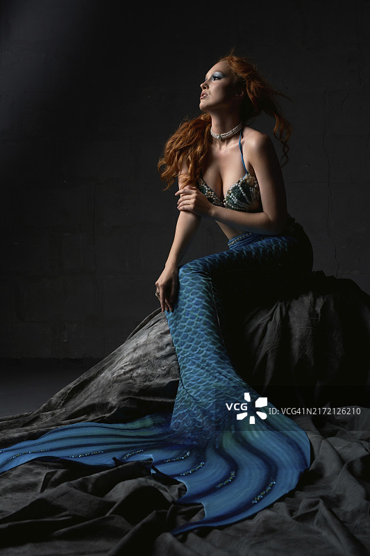 年轻红发女子身穿胸罩和蓝色美人鱼尾坐在岩石上，背靠黑色背景并看向远方图片素材