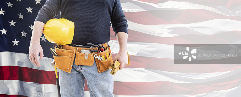 男承包商佩戴工具腰带，手持空白黄色安全帽和手套，背景是挥舞的美国国旗banner图片素材