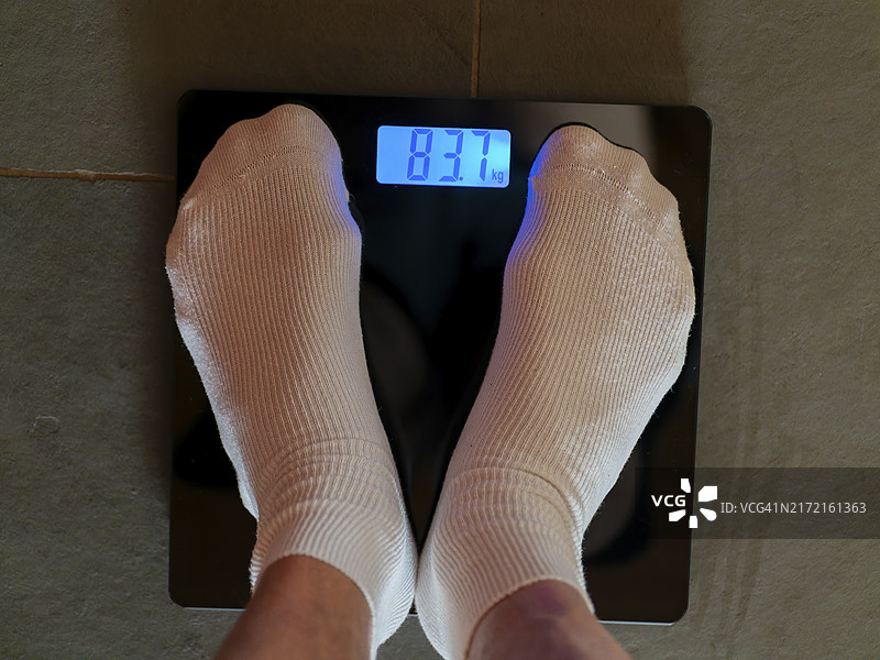 穿着白色袜子站在体重秤上的脚，测量体重，数字体重以公斤显示图片素材
