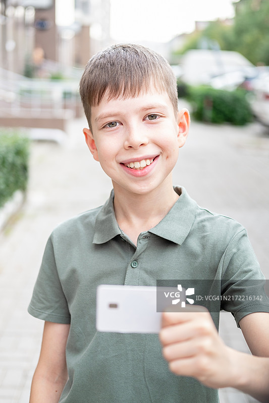 一名12岁男孩穿着绿色T恤，在城市街道上户外拿着一张白色银行卡的肖像图片素材