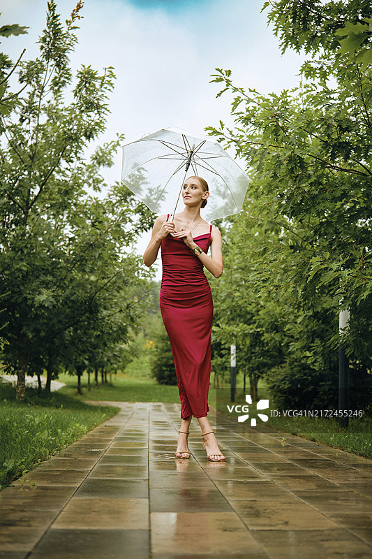 在雨中公园撑伞摆姿势的时尚苗条女子，身穿红色吊带长裙和凉鞋图片素材