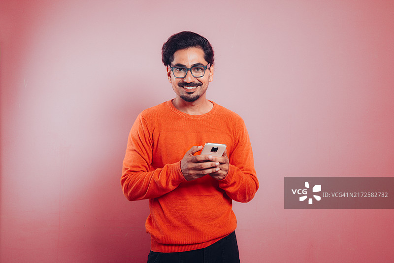 粉色背景前看镜头玩手机的快乐年轻男人图片素材