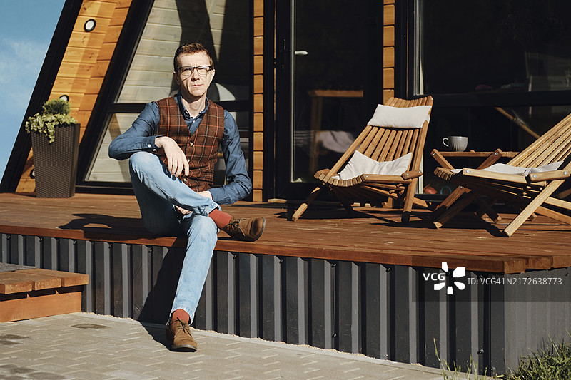清晨坐在木屋露台上，身穿牛仔裤、衬衫和背心的中年男人图片素材