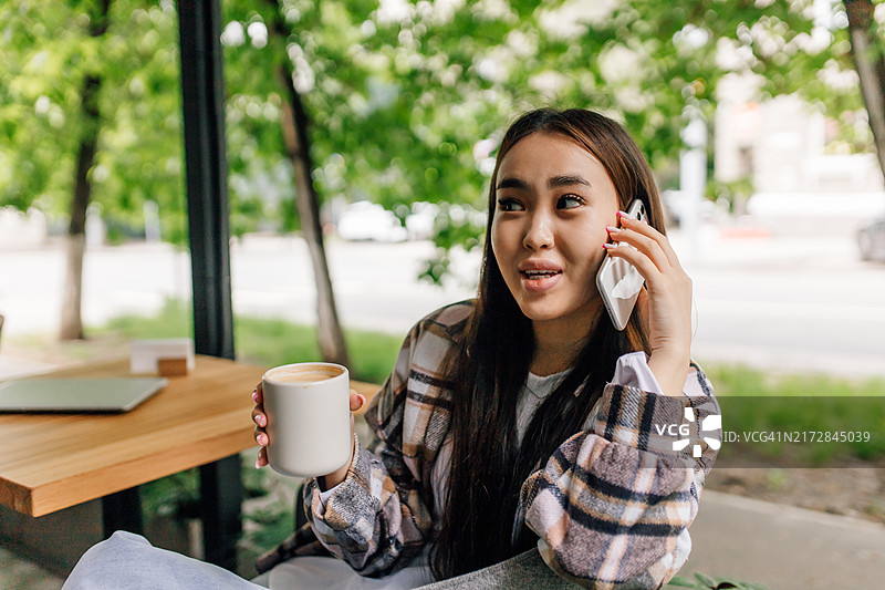 亚洲籍Z世代女商人坐在室外咖啡馆的桌旁，通过电话与客户沟通图片素材