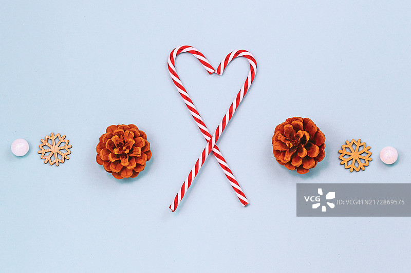 以红色和白色条纹糖果手杖形成心形的季节性冬季平铺，浅蓝色背景上的松果、木制雪花装饰品和白色雪球图片素材