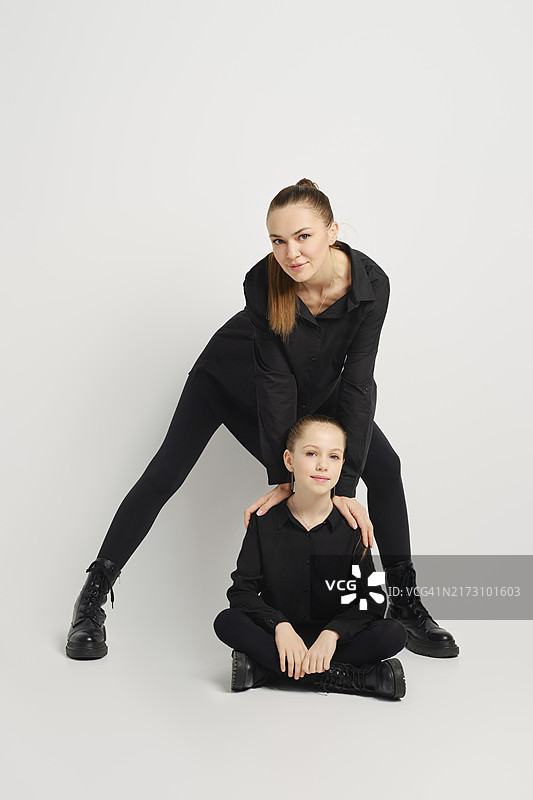 穿着相似黑色服装的现代家庭自信地摆姿势，背景是纯白色。母亲站在坐在地板上的女儿身后，向前倾，将手放在女儿的肩膀上图片素材