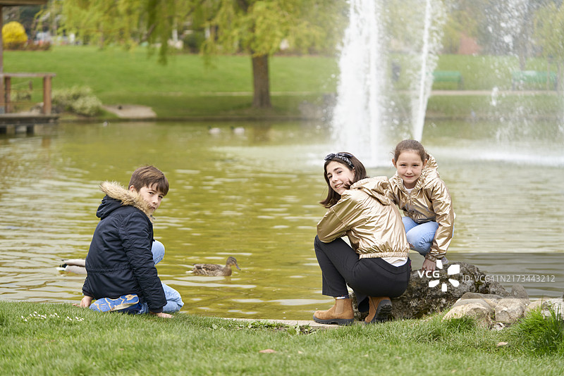 三个穿着外套的男孩坐在公园湖岸边的岩石上，回头对着镜头微笑，后视图图片素材