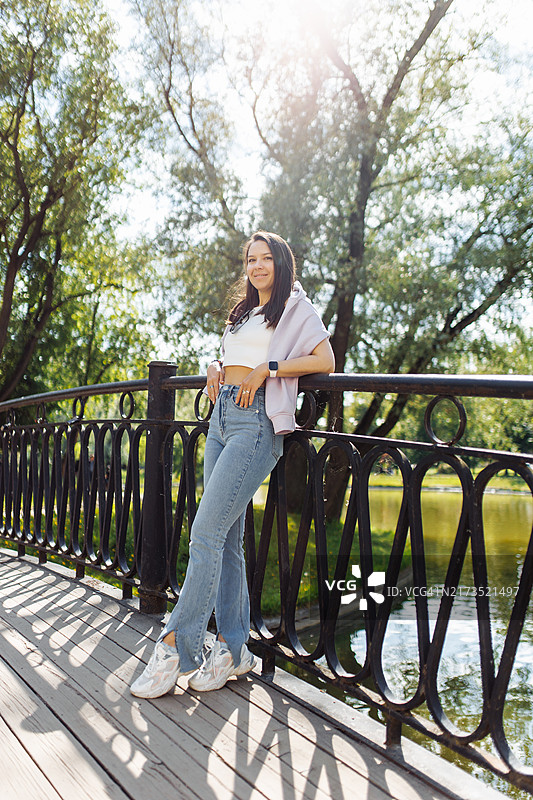 可爱的女人穿着白色背心、牛仔裤和淡紫色毛衣，倚靠在人行天桥的栅栏上图片素材