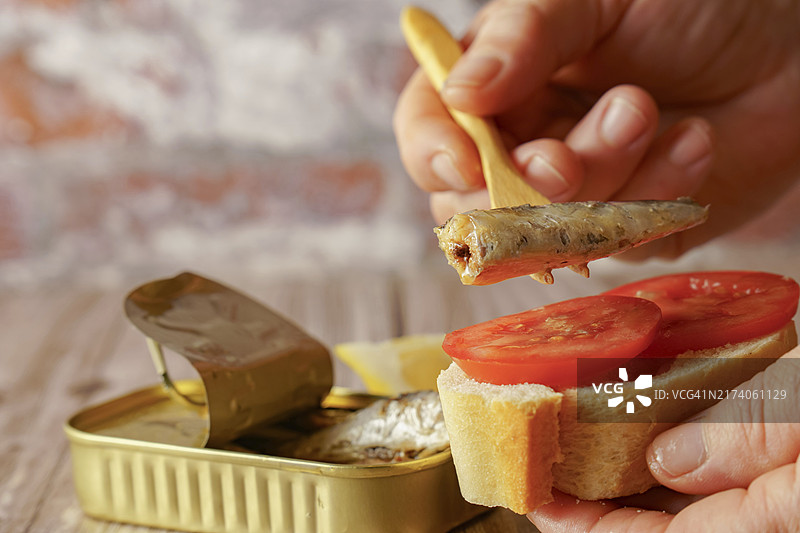 一位女士在面包片上放沙丁鱼，背景是沙丁鱼罐头和木桌上的柠檬图片素材