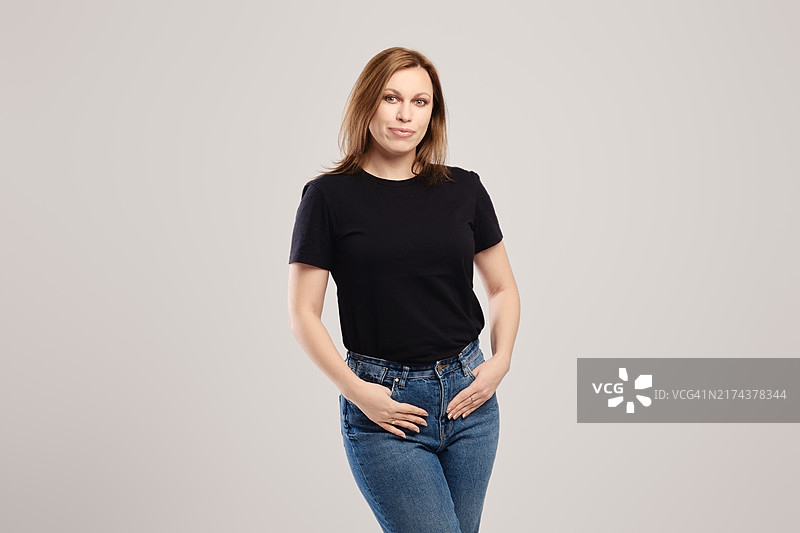 穿黑色T恤和牛仔裤的年轻女人在灰色背景前摆姿势图片素材