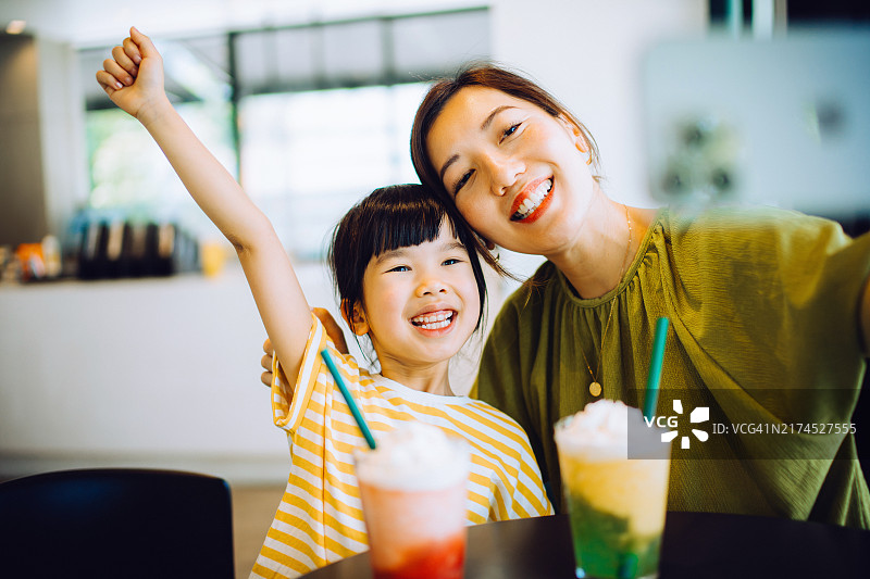 快乐的年轻亚洲母亲和女儿在时尚的咖啡馆里度过一天，用智能手机一起自拍，同时享受清爽的冰镇水果冰沙，母女情，捕捉回忆，家庭爱与团结图片素材