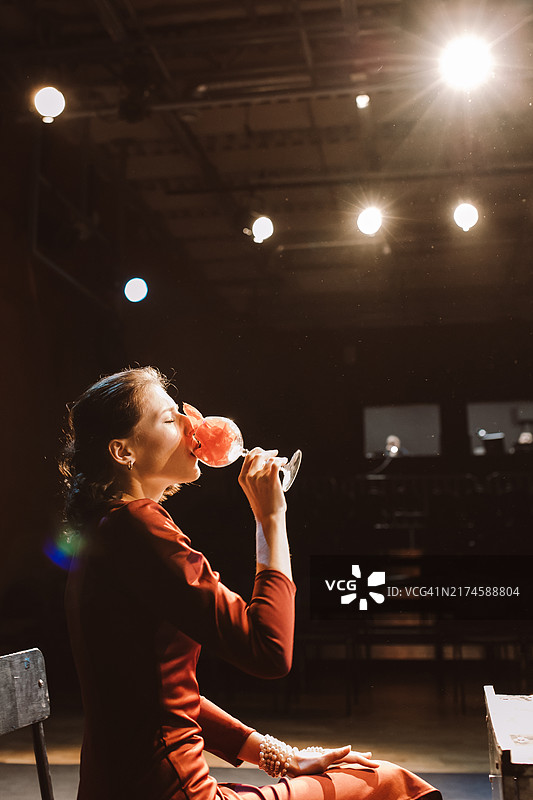 身穿红裙的女子在大学戏剧社舞台表演中手持酒杯坐在椅子上图片素材