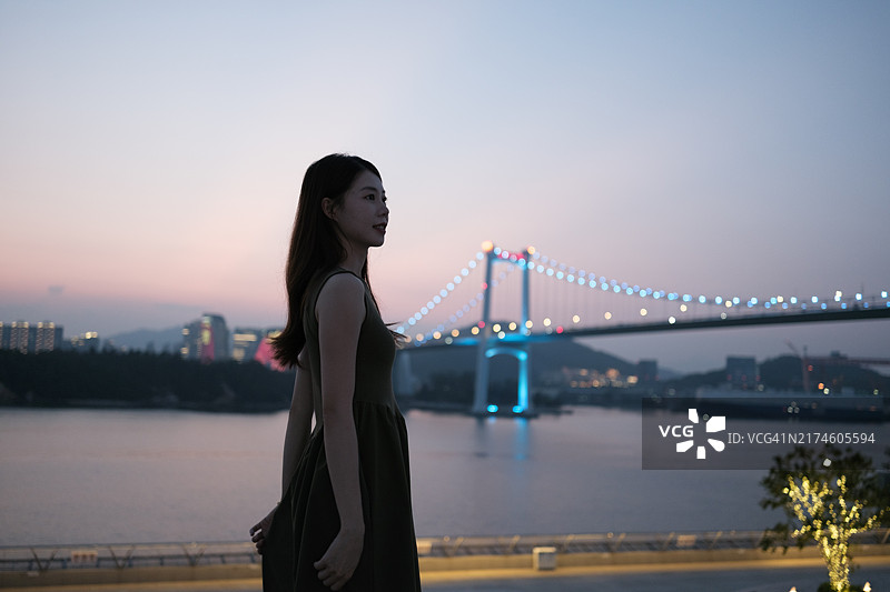 年轻女人站在河岸眺望，背景是宏伟桥梁图片素材