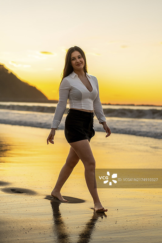 夕阳下，时尚的女性赤脚走在海滩上的竖版肖像图片素材