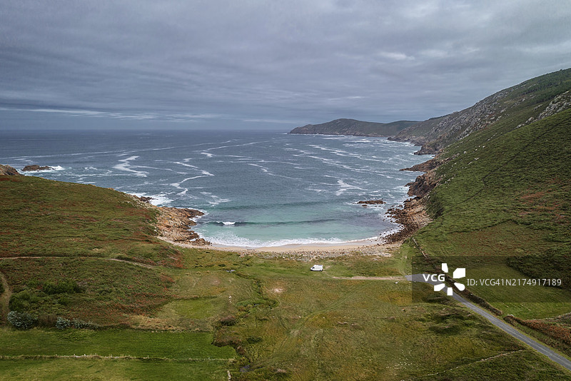 在欧洲西班牙加利西亚的野生海滩上，一辆露营车停靠在绿色景观中的无人机航拍图片素材