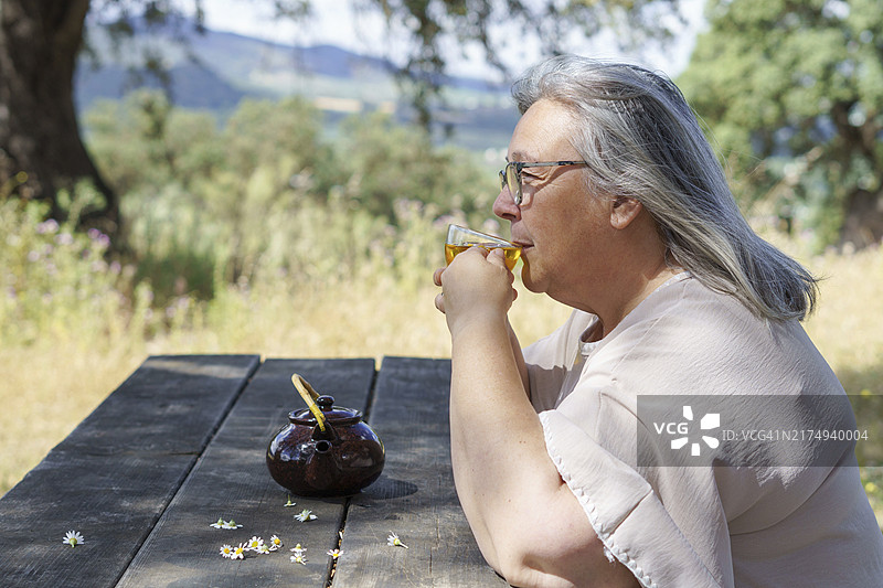 女子在田野木桌上品尝洋甘菊茶的侧面照图片素材