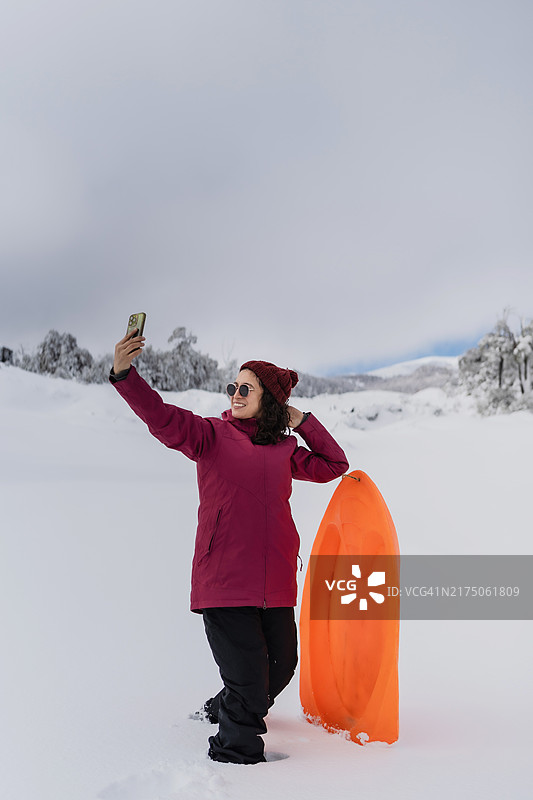 穿着红外套的女人拿着橙色雪橇自拍图片素材