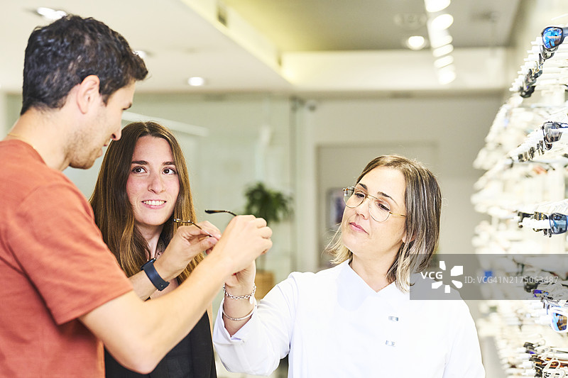 年轻夫妇站在眼镜店里与医生交谈并向她展示眼镜的侧视图图片素材