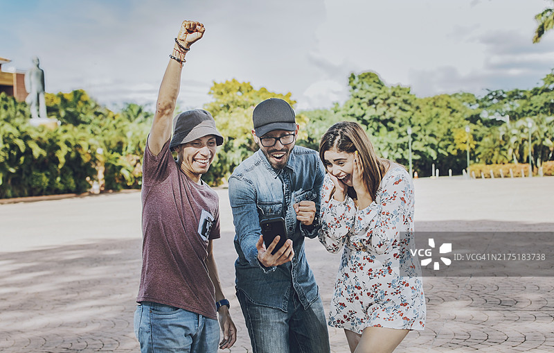 三个快乐的人在户外看手机，兴奋的青少年朋友在街上看手机屏幕图片素材