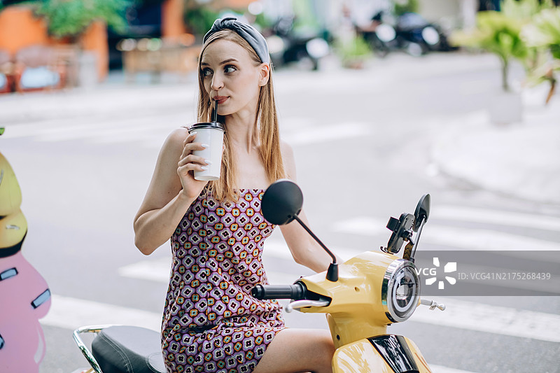 年轻的金发女性穿着印花裙，坐在黄色滑板车上喝外卖杯里的饮料。背景是城市道路，带有运动模糊效果。图片素材