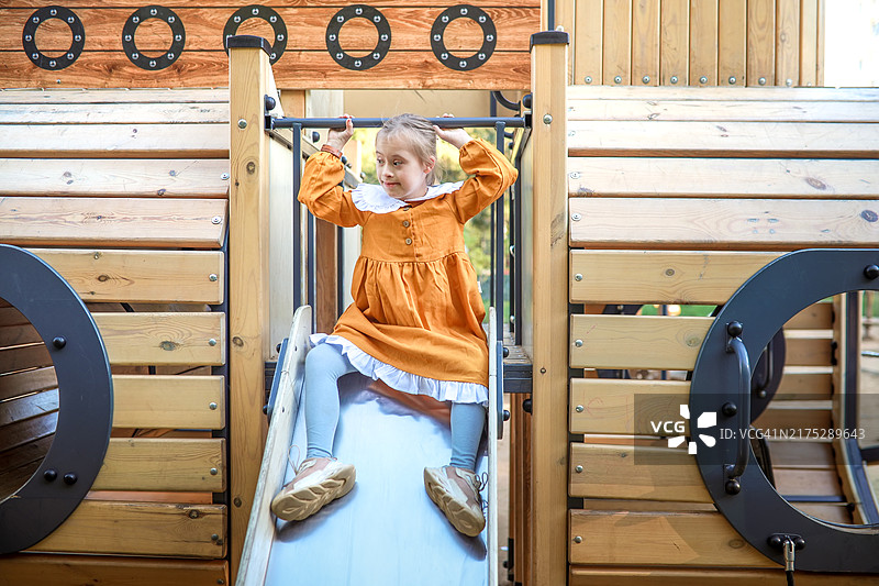 穿着芥末色连衣裙的唐氏综合征快乐女孩在夏天玩滑梯图片素材