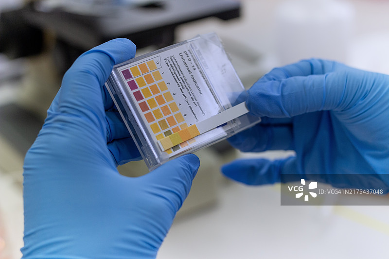 戴着手套的手拿着尿液试纸测试盒。测试盒显示了一个颜色图表，用于比较尿液分析的pH值结果。图片素材