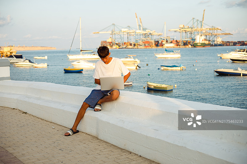 少年坐在码头上使用笔记本电脑，远处是宁静的海面和起重机图片素材