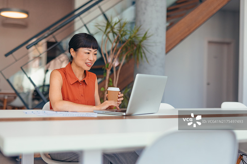 女人在现代办公室一边喝咖啡一边用笔记本电脑工作图片素材