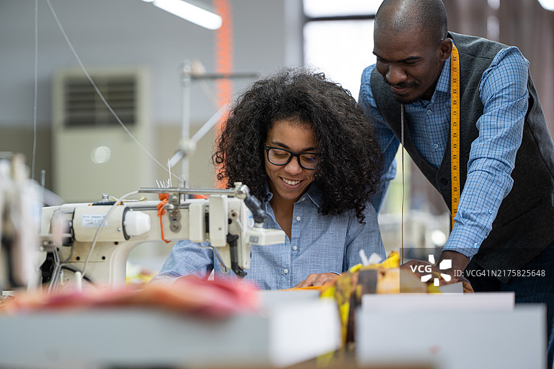 非洲裔美国年轻同事在服装制造工作室讨论，女人在现代缝纫机上缝纫图片素材