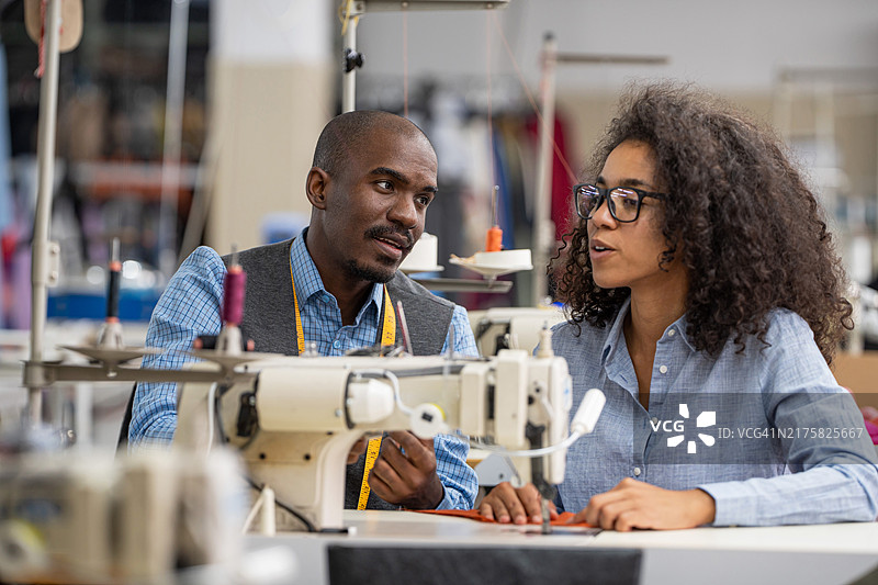 非洲裔美国年轻同事在服装制造工作室讨论，女人在现代缝纫机上缝纫图片素材
