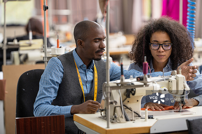 非洲裔美国年轻同事在服装制造工作室讨论，女人在现代缝纫机上缝纫图片素材