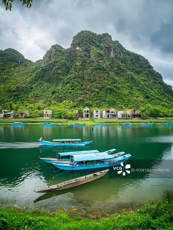 峰牙-己榜国家公园：喀斯特山脉森林和传统房屋在水中的倒影，一艘蓝色小木船在河上行驶（越南广平省）图片素材