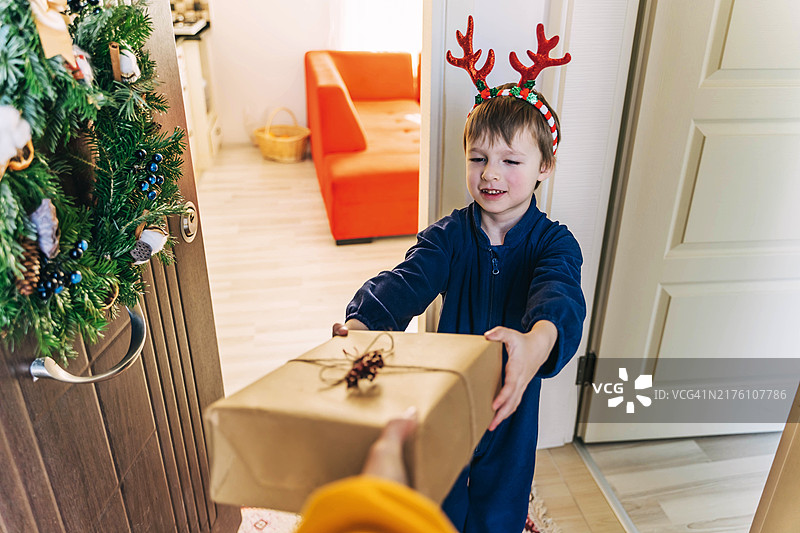 穿睡衣戴鹿角的小男孩收到圣诞礼物，门上挂着圣诞花环图片素材