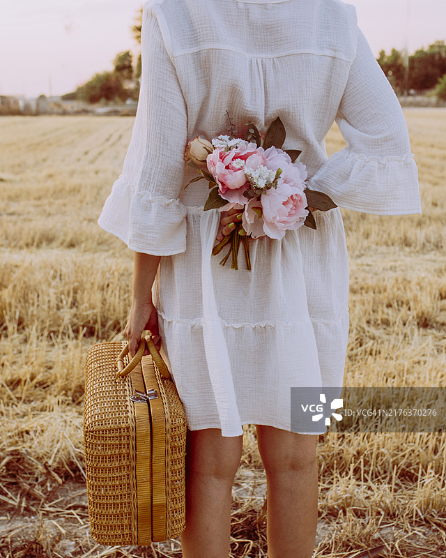 背影：女性在麦田日落时分手持野餐篮和花束图片素材