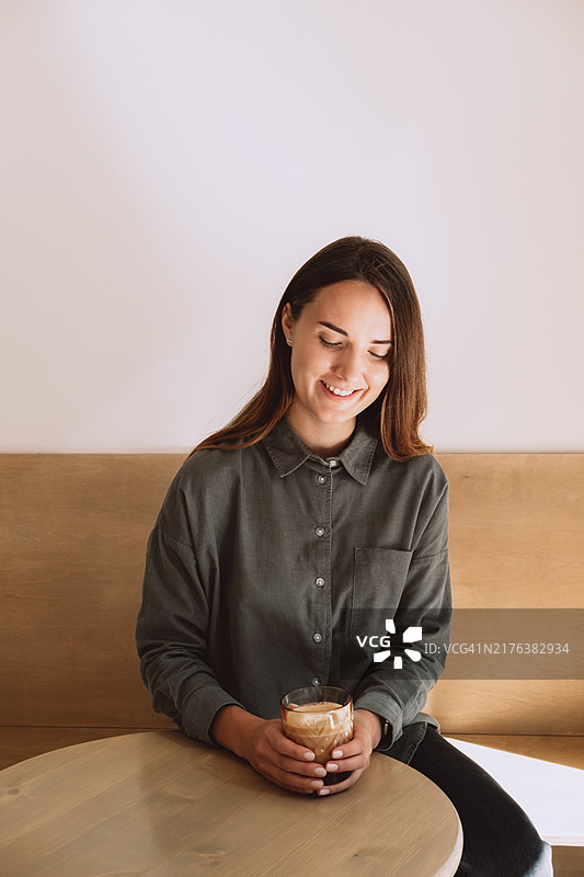 身穿绿色亚麻衬衫的年轻女人在咖啡店的木桌旁喝拿铁咖啡图片素材