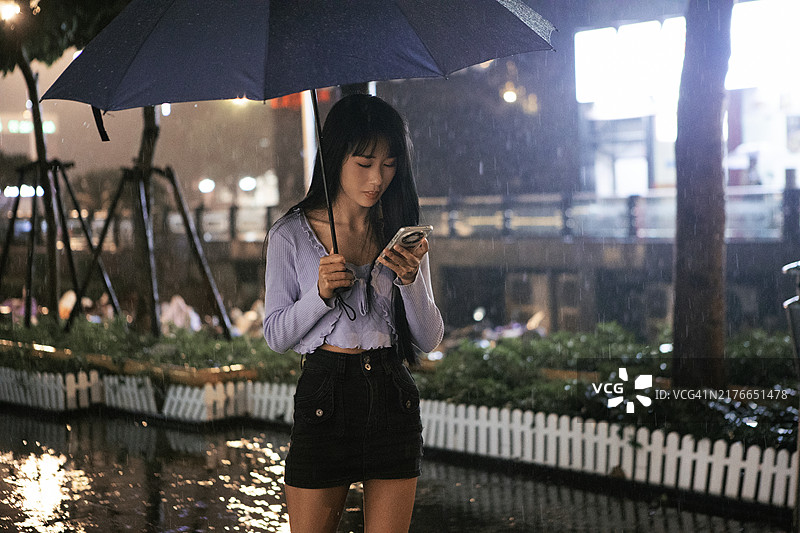 夜晚，一位亚洲女人在城市里撑着伞，拿着手机图片素材