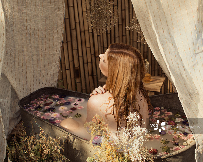 一位红发女性正在花瓣满溢的浴缸中沐浴，浴缸位于花园里。图片素材
